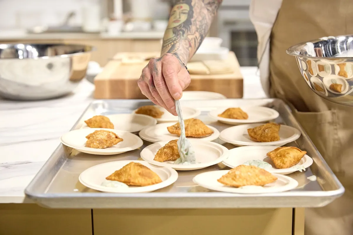 Chef with tattooed arm plating golden fried samosas with yogurt sauce on small dishes