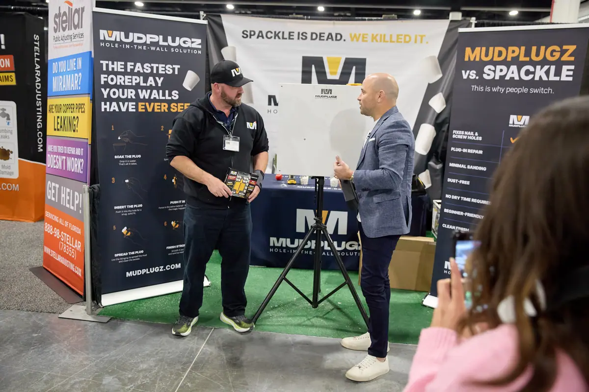 Mudplugz exhibitor demonstrates wall-repair product to a visitor at a home show booth