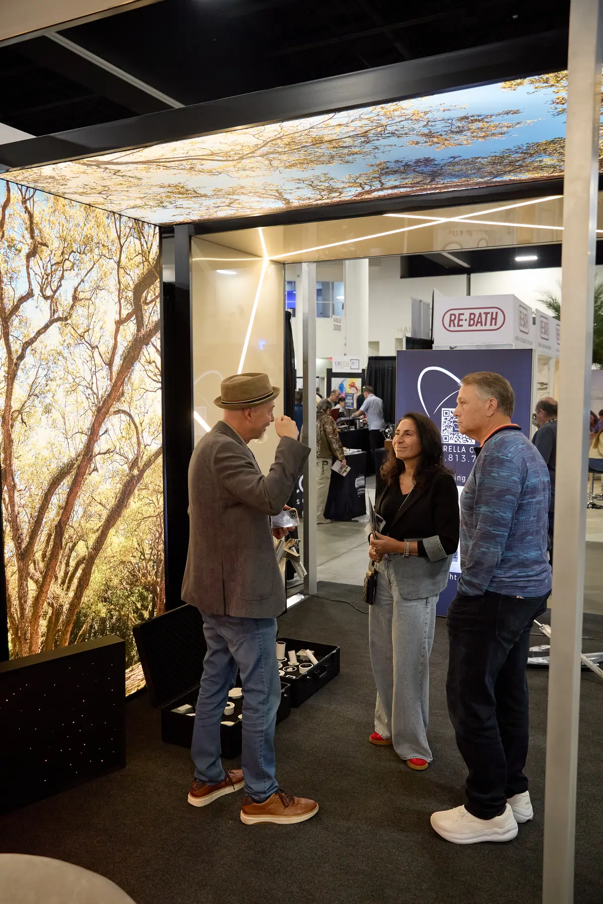 Three visitors chat inside a booth featuring large-format nature photo wall panels and a Re-Bath sign