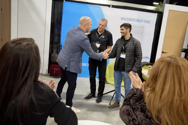 Two men shake hands at a door exhibitor booth while a third man looks on and audience claps