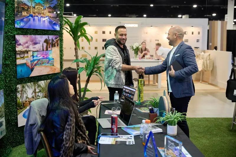 Exhibitor shakes hands with a visitor at a pool and outdoor living booth with tropical greenery backdrop