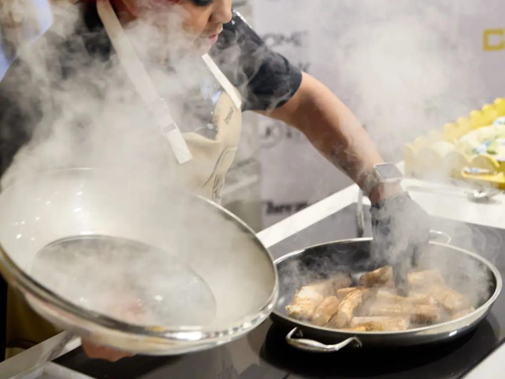 Chef in a white apron sears fish in a steaming skillet at an exhibitor culinary stage