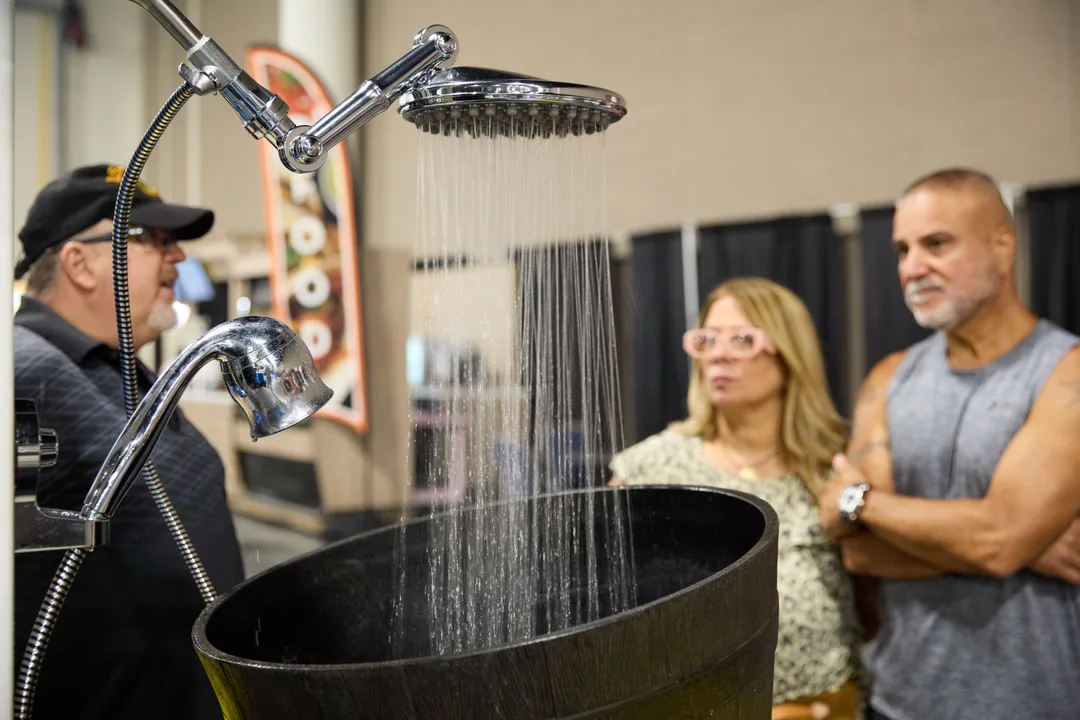 Close-up of chrome rainfall and handheld shower heads spraying water into a black basin with a couple watching the demo at a spa-shower booth.