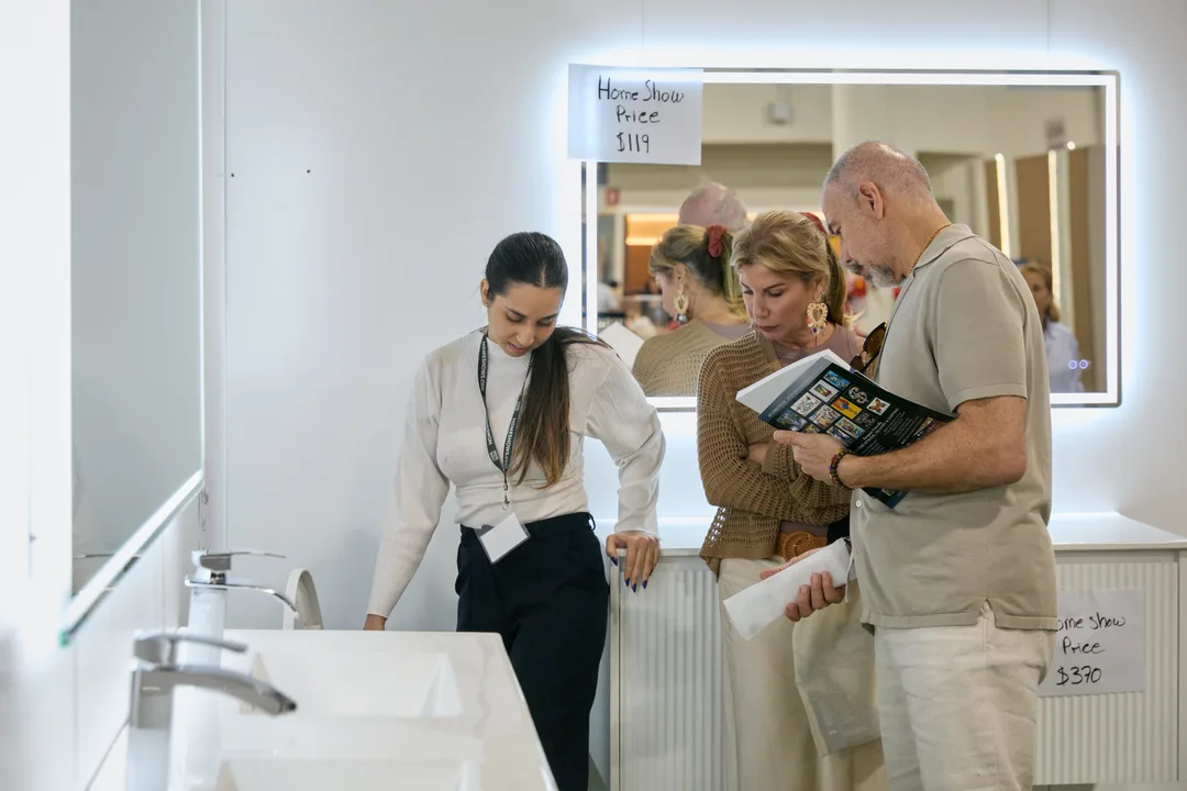 A woman in a white blouse shows white vanity sinks to a couple browsing a sample catalog at a bathroom vanity booth with backlit mirrors and printed signage, landscape orientation.