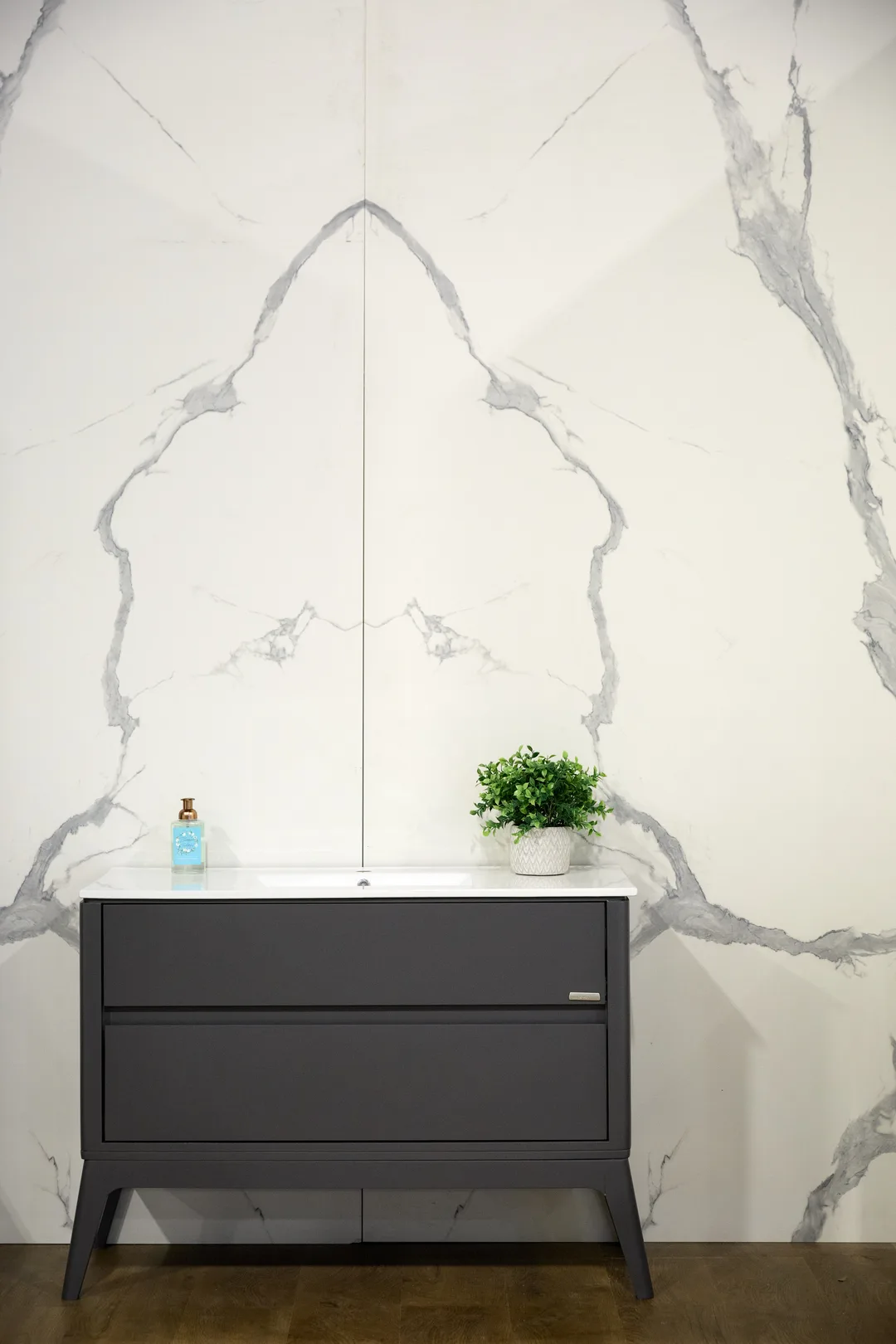 A dark gray modern bathroom vanity with white sink sits against a bookmatched white-and-gray marble-look slab wall with a small green plant, in a portrait interior detail shot.
