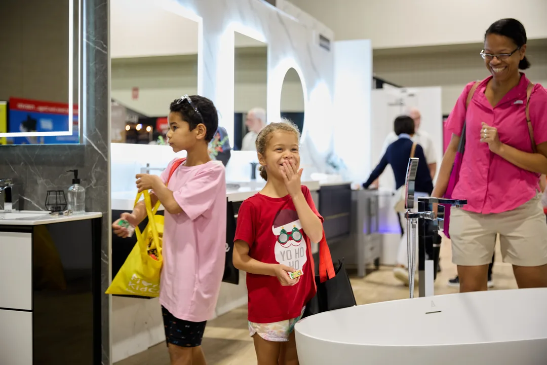 Two children, one giggling with a hand over her mouth, stand by a white freestanding tub in a bath showroom booth as a staffer in pink walks up, landscape orientation.
