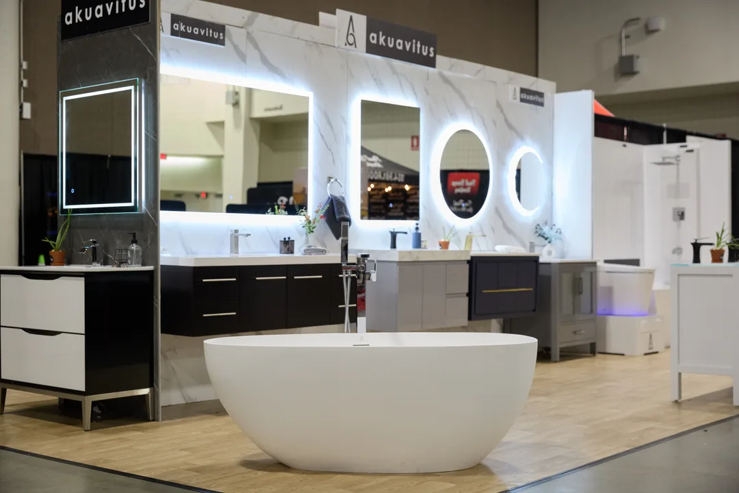 The Akuavitus bathroom booth centers on a white oval freestanding soaking tub in front of a wall of LED mirrors and vanities with marble-look backdrop, in a landscape show-floor shot.