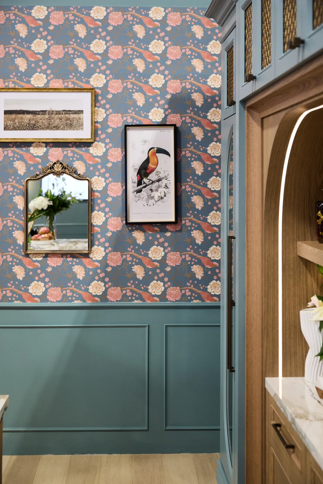 A teal-panelled powder room with floral-and-toucan wallpaper, framed bird and landscape prints, a gold-framed mirror and an arched marble niche, portrait orientation.