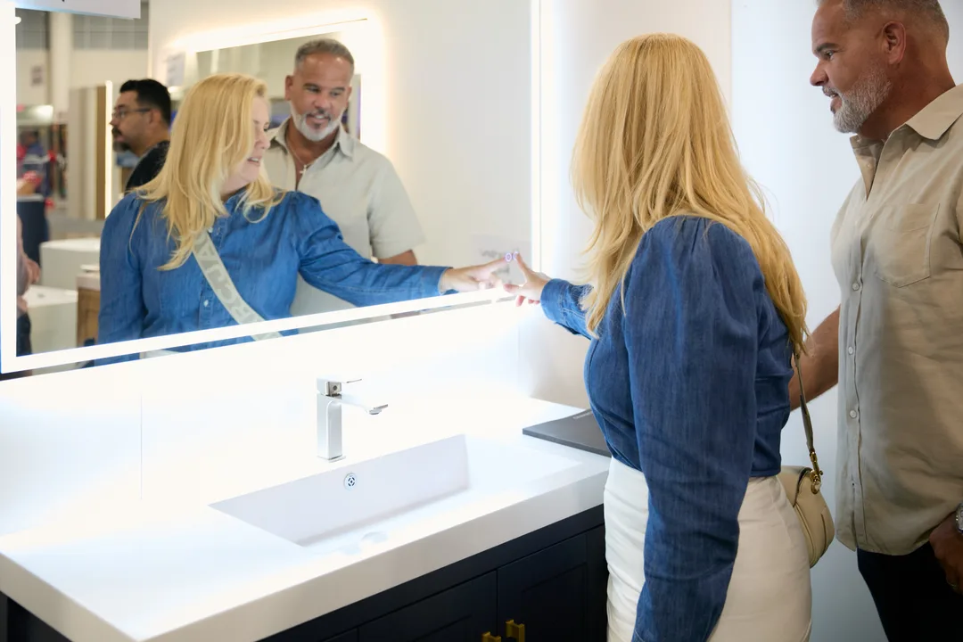 A couple stands at a backlit bathroom mirror over a white vanity while a vendor's reflection is visible in the glass at a bath booth, landscape.