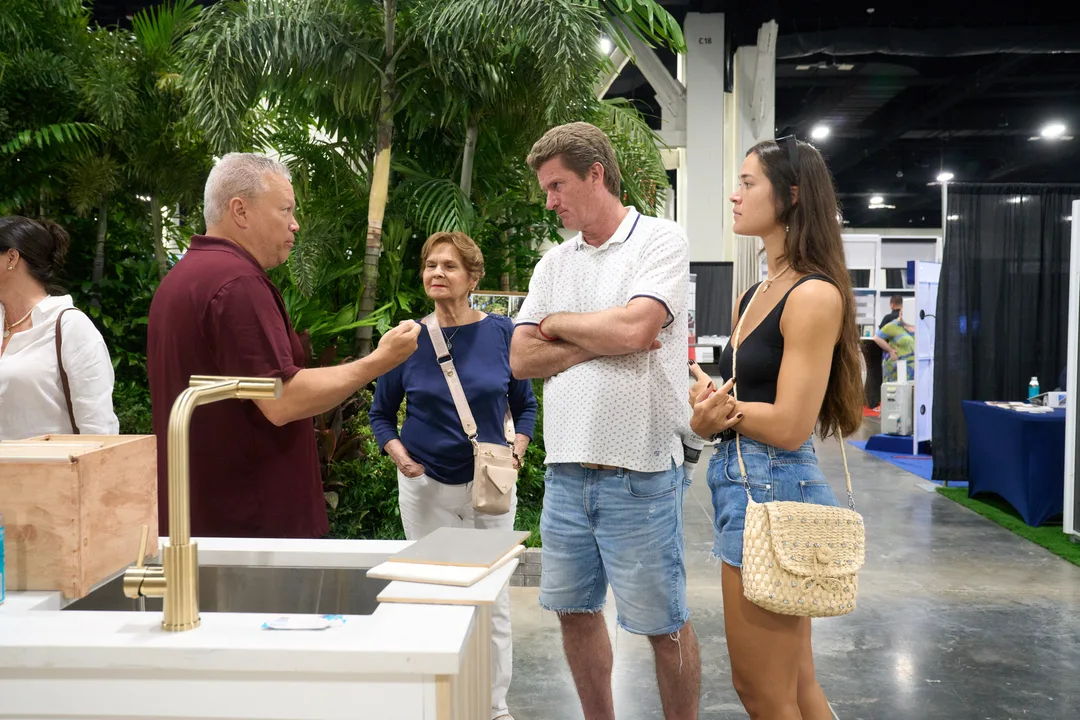 A man in a maroon polo demonstrates a brass faucet on a white sink vanity to attentive visitors at a tropical-themed kitchen-and-bath booth, landscape orientation.