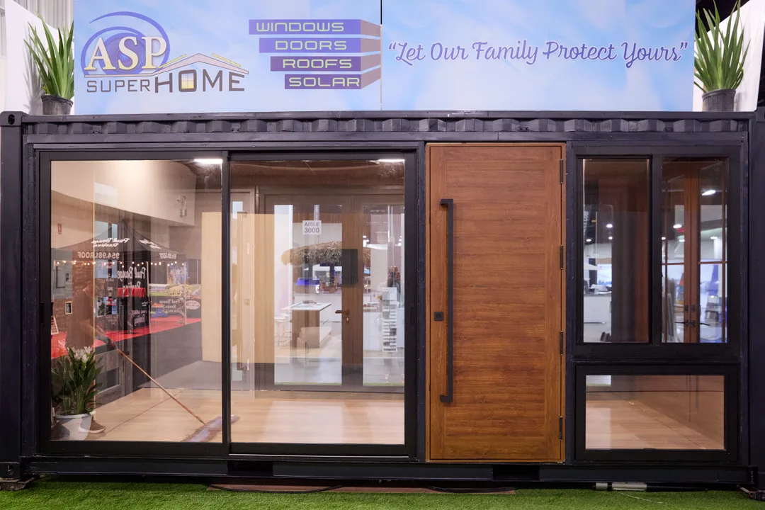 The ASP SuperHome shipping-container booth shows a glass slider and wood pivot front door under the tagline "Let Our Family Protect Yours" with windows-doors-roofs-solar signage, in a landscape show-floor shot.
