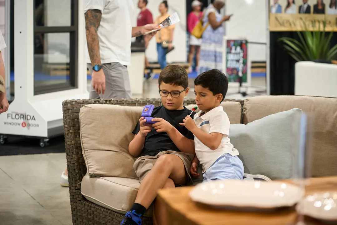 Two young boys sit on a wicker patio sofa engaged with a toy near a Florida Window & Door booth on the show floor, landscape orientation.