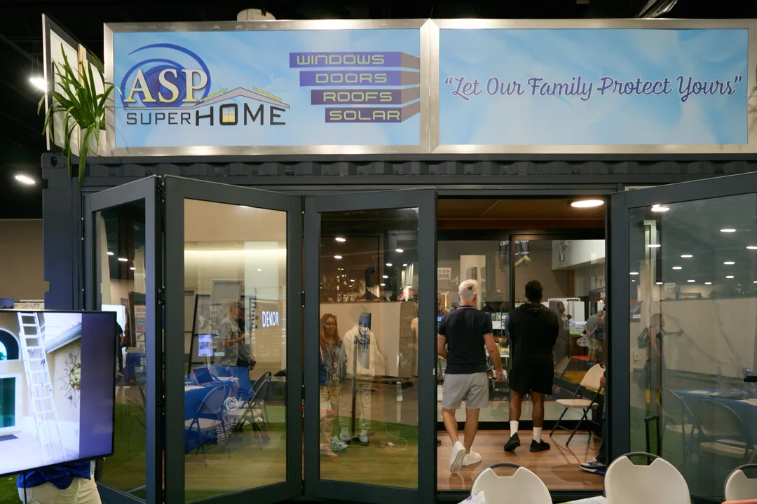Front view of the ASP Super Home shipping-container booth with a sign reading 'WINDOWS DOORS ROOFS SOLAR — Let Our Family Protect Yours' and folding glass doors, with attendees inside, landscape orientation.