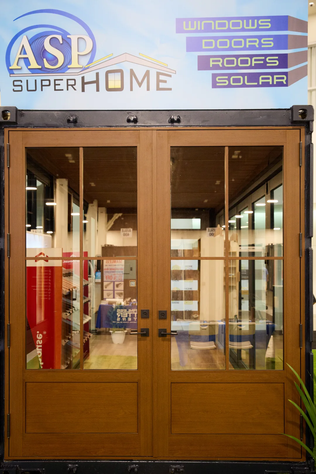A double wood-and-glass French door set into a black shipping-container booth branded ASP SuperHome with WINDOWS, DOORS, ROOFS, SOLAR signage, in a portrait show-floor shot.