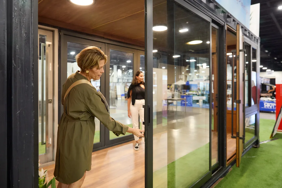 A woman in an olive dress slides open a large black-framed glass door on a modular home display while another woman stands inside, on green turf flooring, landscape orientation.