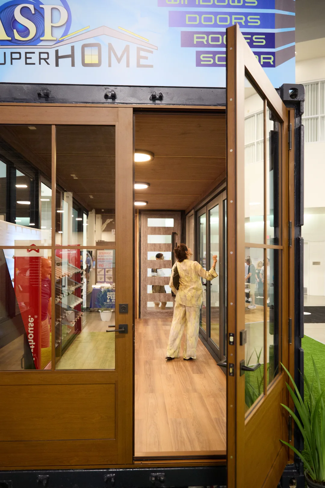 A woman with a crossbody bag opens an interior glass door inside the wood-lined ASP Super Home container house display, portrait orientation.