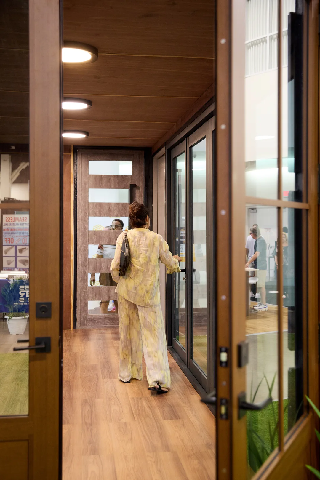 A woman steps through the open glass doors into the wood-paneled ASP Super Home modular home booth promoting doors, roofs and solar, portrait orientation.