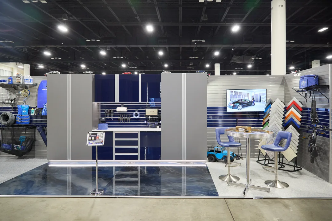 A blue-and-grey modular garage-organization booth showing slatwall cabinetry, tool storage, a wall-mounted display screen, garage flooring tiles and two purple bar stools at a high table, landscape orientation.