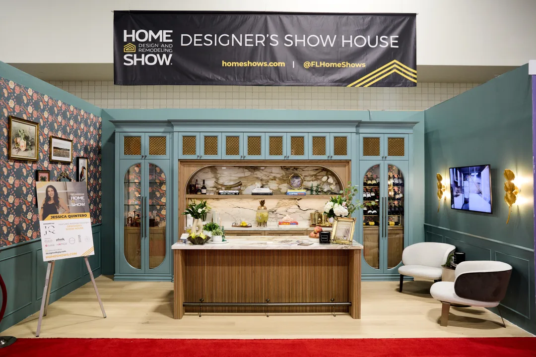 A wide view of a teal Designer&rsquo;s Show House booth: cane-front glass upper cabinets flanking an arched marble-backed bar niche, a fluted wood bar front, floral wallpaper and gold leaf sconces, with two curved lounge chairs at right, landscape orientation.