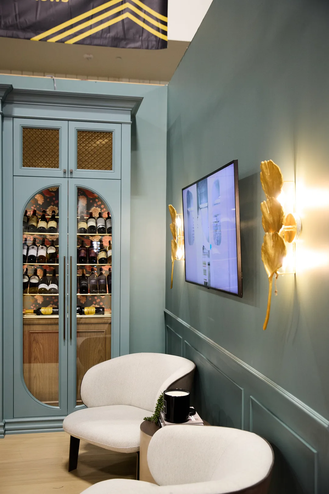 A teal-walled lounge nook with an arched glass wine cabinet, wall-mounted TV, gold leaf sconces and two curved white accent chairs, portrait orientation.