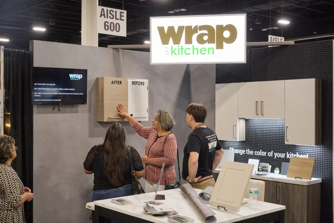 Visitors examine before-and-after cabinet door samples at the 'Wrap My Kitchen' booth with a marble-topped table of materials under Aisle 600 signage, landscape orientation.
