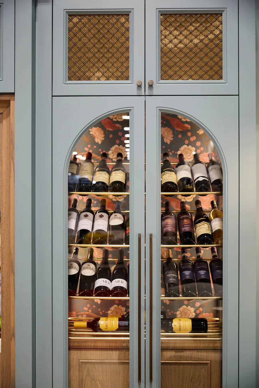 A built-in sage-green cabinet with arched glass doors and brass mesh panels displaying rows of wine bottles against floral wallpaper, portrait orientation.