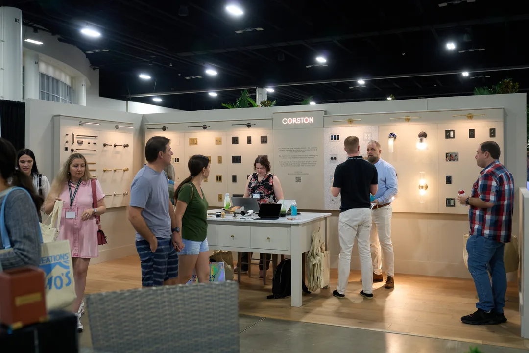 Visitors gather around a white desk in the Corston booth, which displays rows of cabinet hardware and lighting on white walls, landscape orientation.