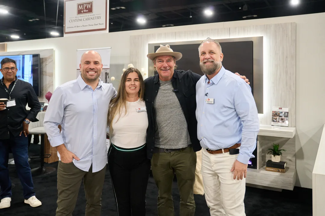 A group portrait of four people smiling together at the MPW Custom Cabinetry booth media wall, the man in the tan hat at center, landscape orientation.