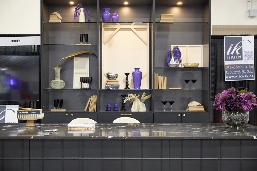 An iKitchen booth shows a black fluted kitchen island with marble-look top and white chairs in front of a styled black shelving wall with purple decor, set on a cowhide rug, in a portrait show-floor shot.
