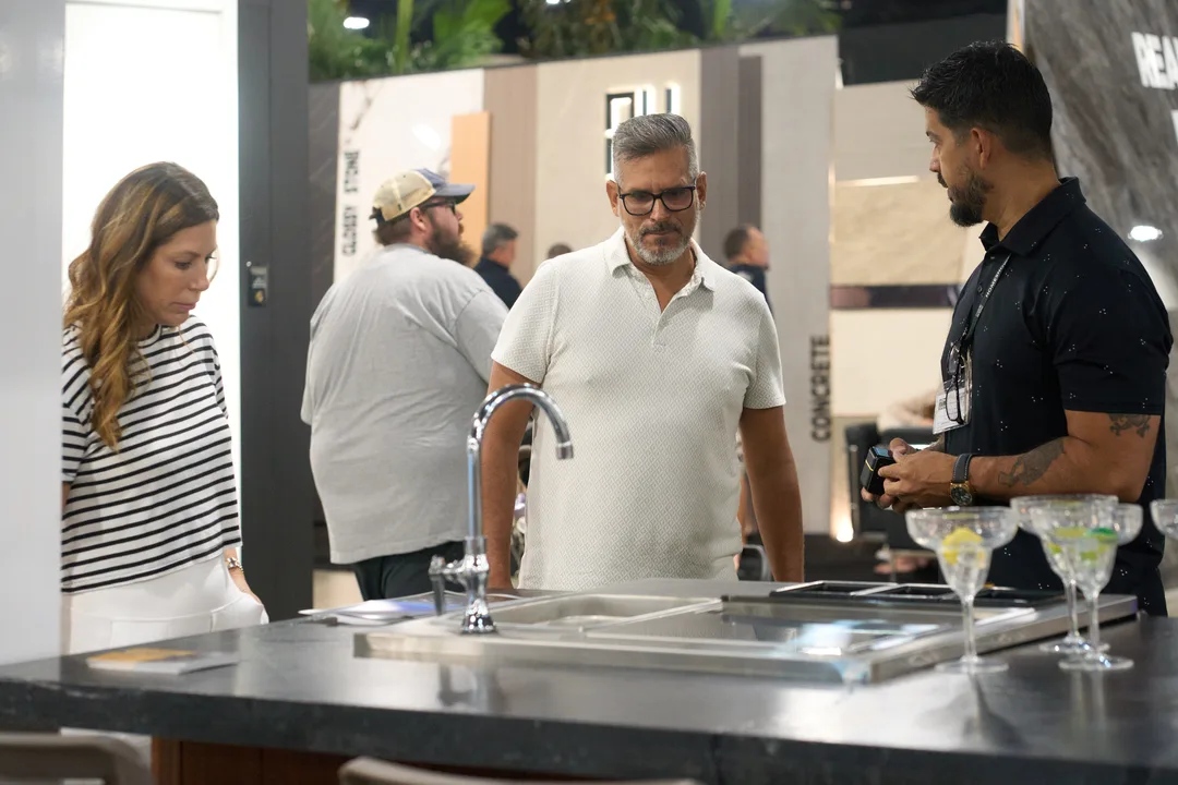A booth staffer in a black shirt shows a black sink-and-faucet countertop display to a woman in a striped top and a man in a white polo at a 'Concreti' stone booth.
