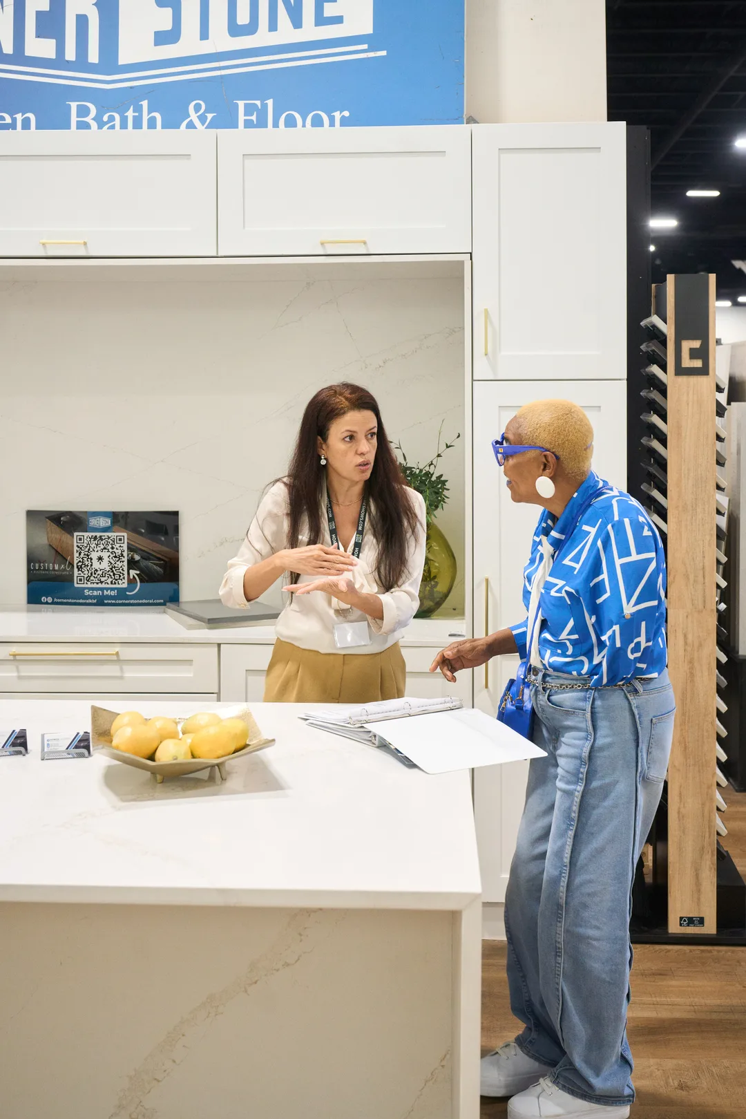 A booth representative and a customer in a blue patterned shirt converse across a white quartz kitchen island with a bowl of lemons at the Corner Stone Kitchen, Bath & Floor booth.