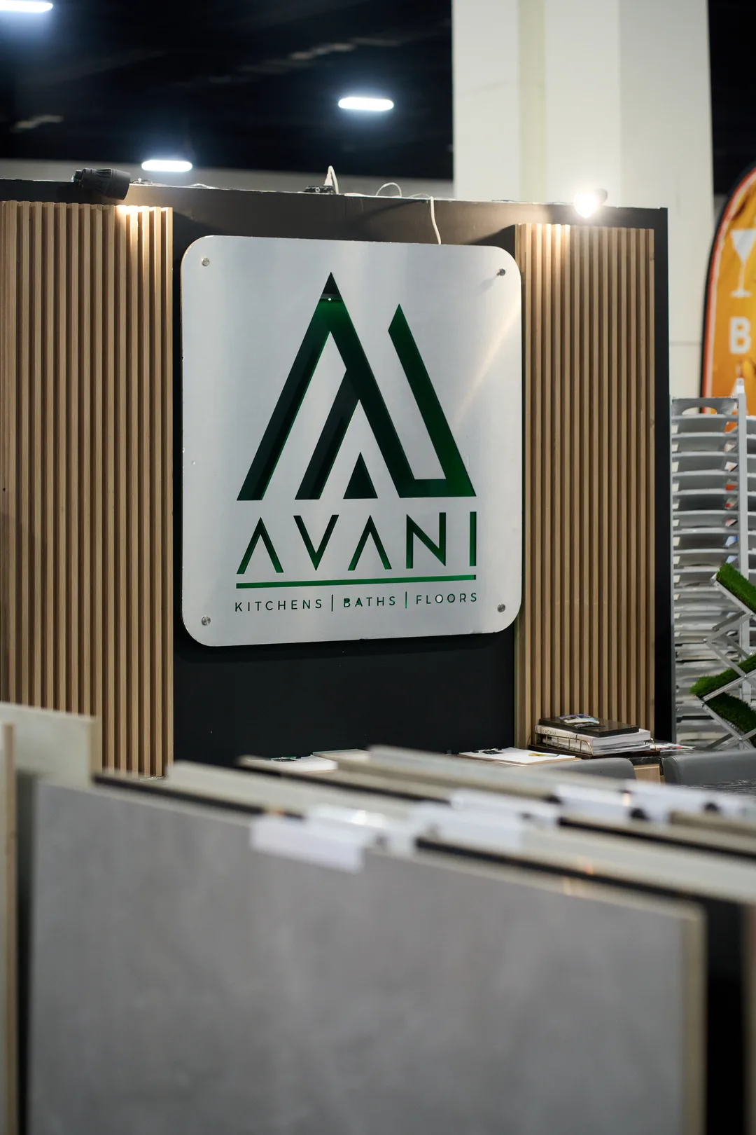 A white 'AVANI Kitchens | Baths | Floors' booth sign with a green logo, mounted on wood-slat panels above racks of large-format tile and stone samples.