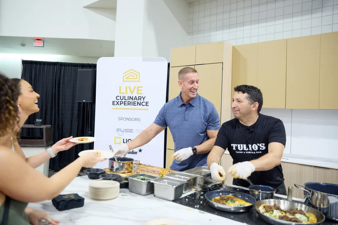 Two chefs serve tasting plates to a woman over the demo kitchen counter in front of a 'Live Culinary Experience' banner, in a candid show-floor scene.
