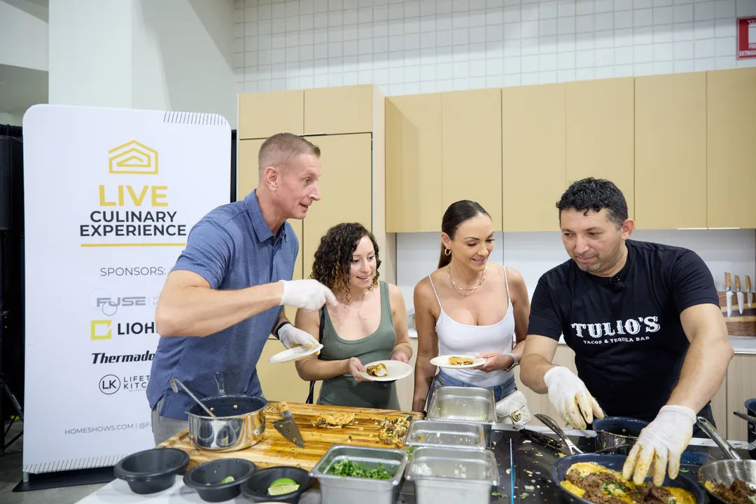 Four people plate food at a kitchen demo counter beside a 'Live Culinary Experience' sponsor banner, with one man in a black Tulio's shirt serving.