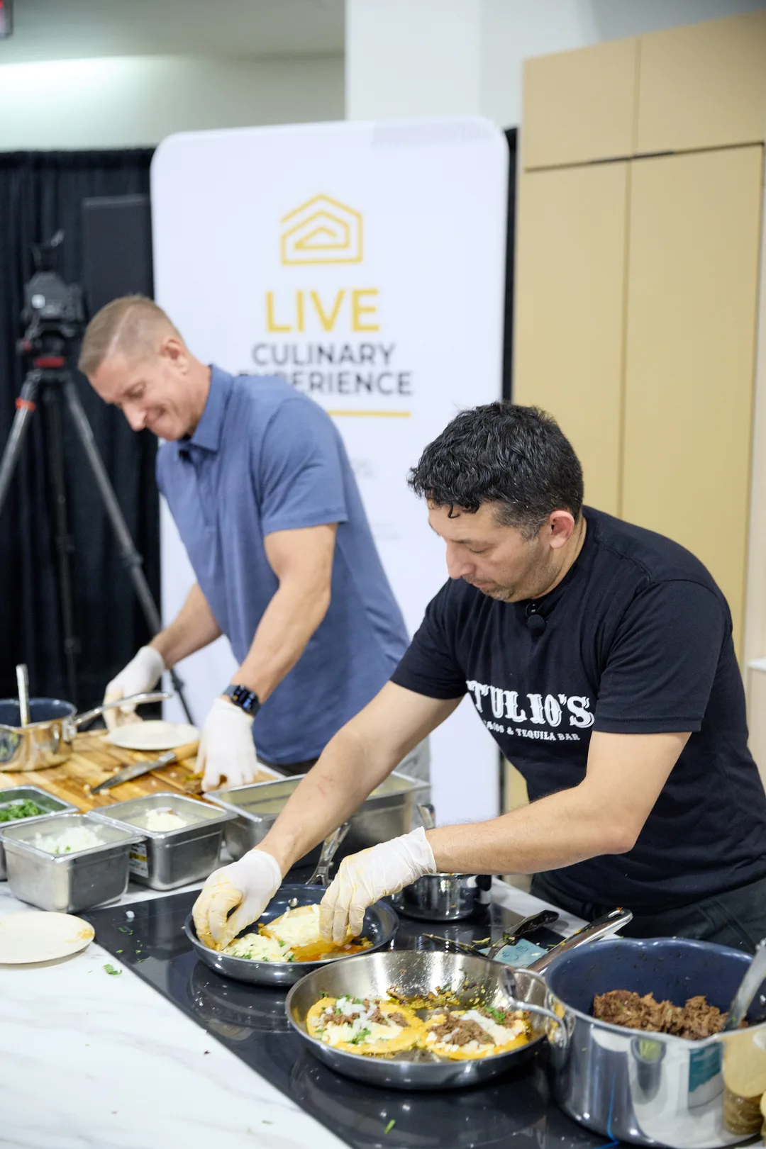 Two chefs assemble tacos and plate food on a white marble demo-kitchen counter crowded with hot pans and prep trays, in front of a 'Live Culinary Experience' banner.