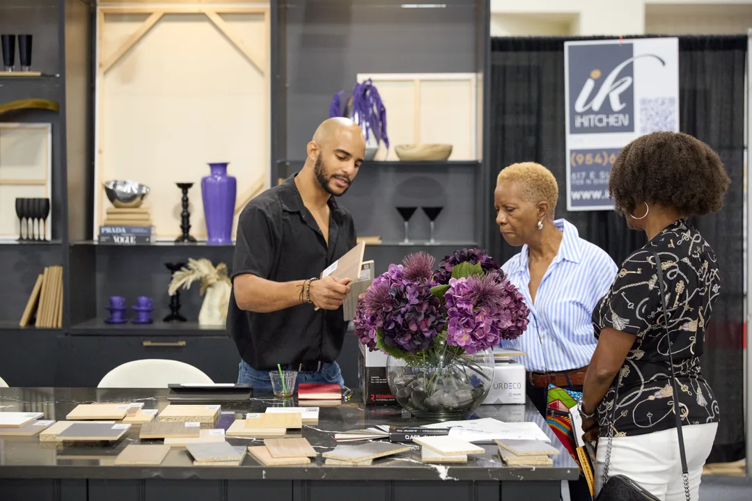 A male iKitchen rep holds up a sample board for two female shoppers at a dark kitchen-island display with purple flowers and dark cabinetry behind.