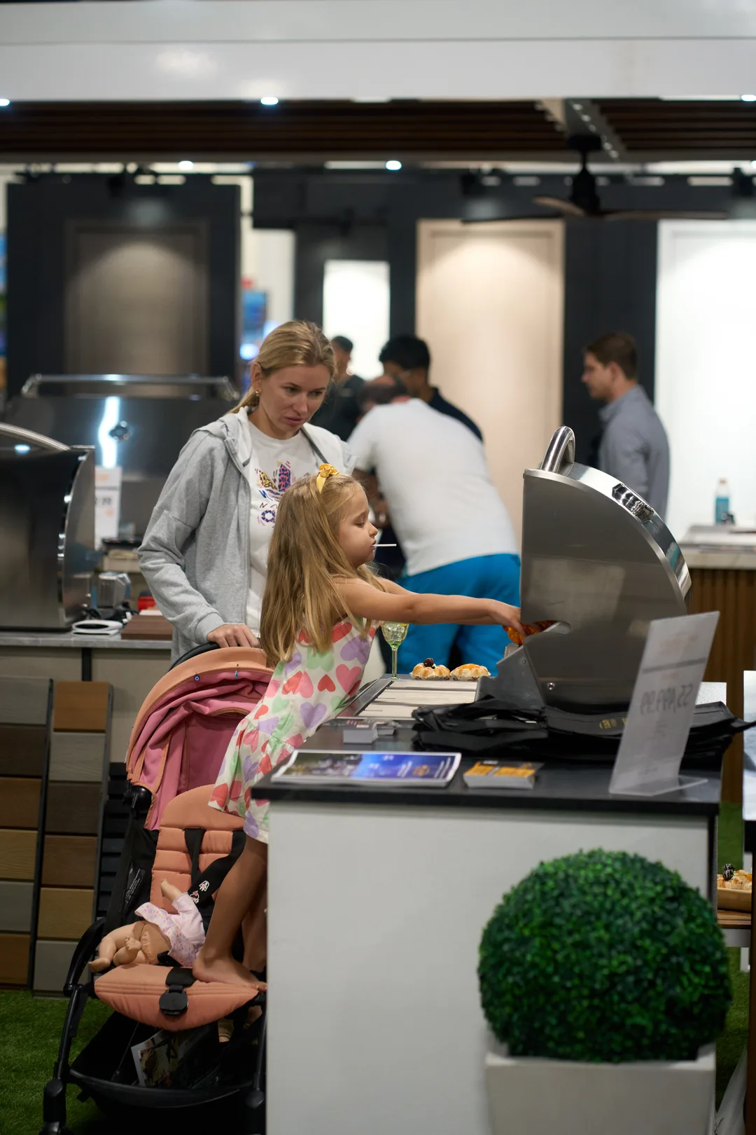 A young girl in a heart-print dress reaches toward a stainless steel grill at an outdoor kitchen booth while a woman watches and a pink stroller sits in front.
