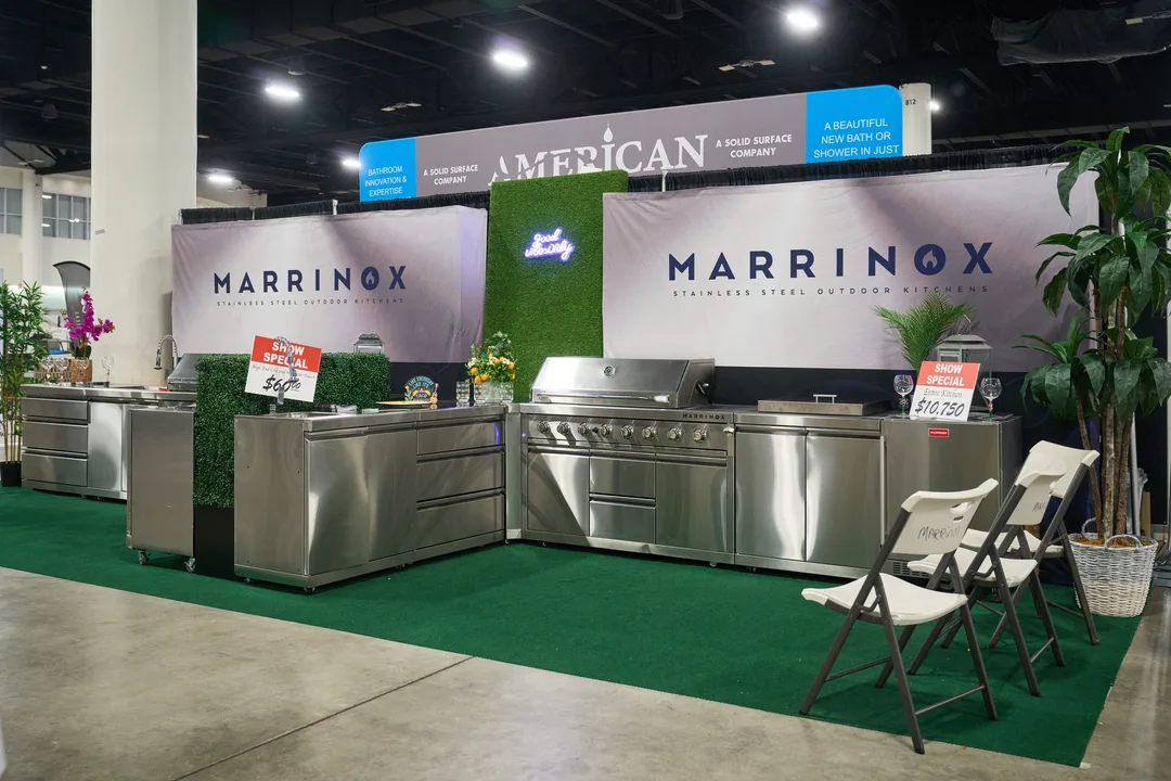 A Marrinox stainless-steel outdoor kitchen booth showing built-in grills and cabinetry on green turf flooring under large brand banners.