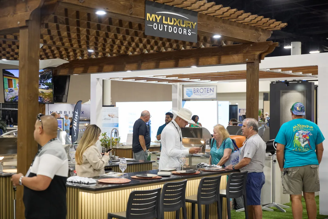 A chef in white demonstrates a cooking station at the wood-pergola My Luxury Outdoors booth as attendees watch from a bar counter on the show floor.