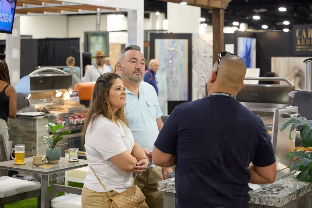 A couple talks with a salesman at an outdoor kitchen booth showing stainless grills and stone countertops, with drinks on the counter.