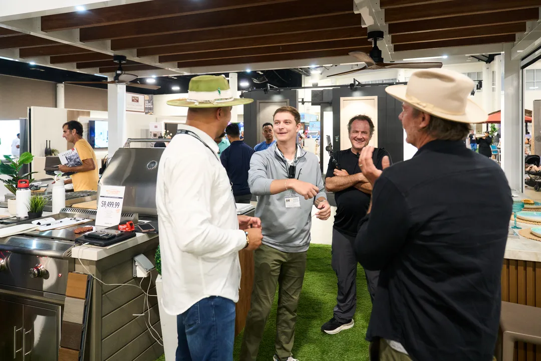 Men chatting and laughing at an outdoor-kitchen grill display booth with artificial turf flooring, two of them in brimmed hats.