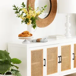 White sideboard with rattan cane doors, decorative mirror, and floral arrangement on top