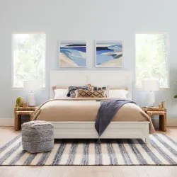 Coastal bedroom with white panel bed, blue seascape artwork, striped rug, and rattan nightstands