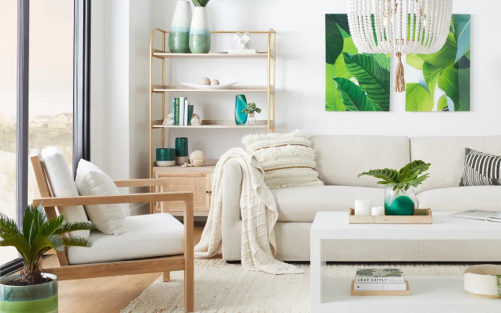 Tropical-inspired living room with white sofa, wood-frame chair, gold shelving, and large leaf canvas art