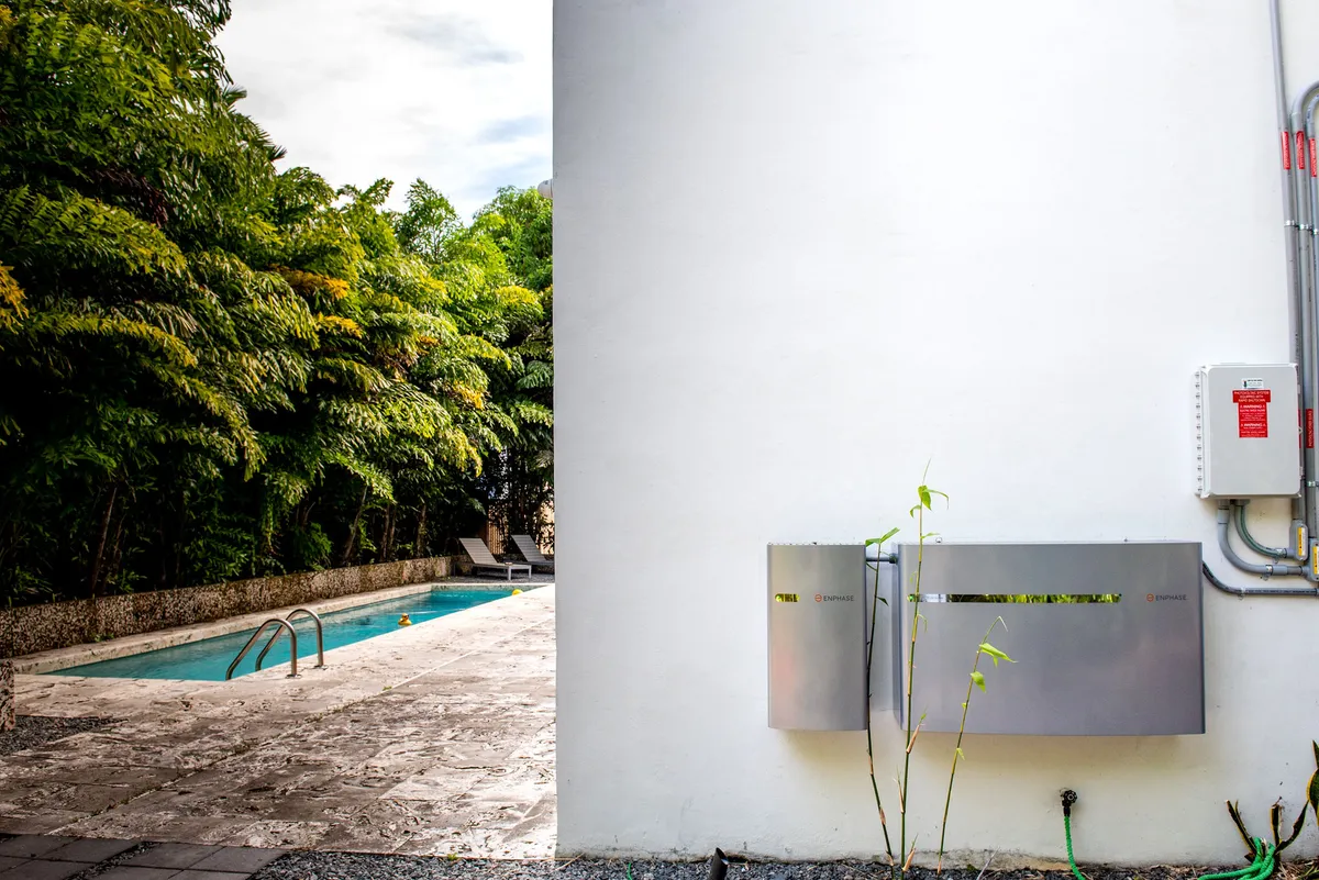 Enphase battery storage units mounted on exterior wall beside backyard pool with tropical trees