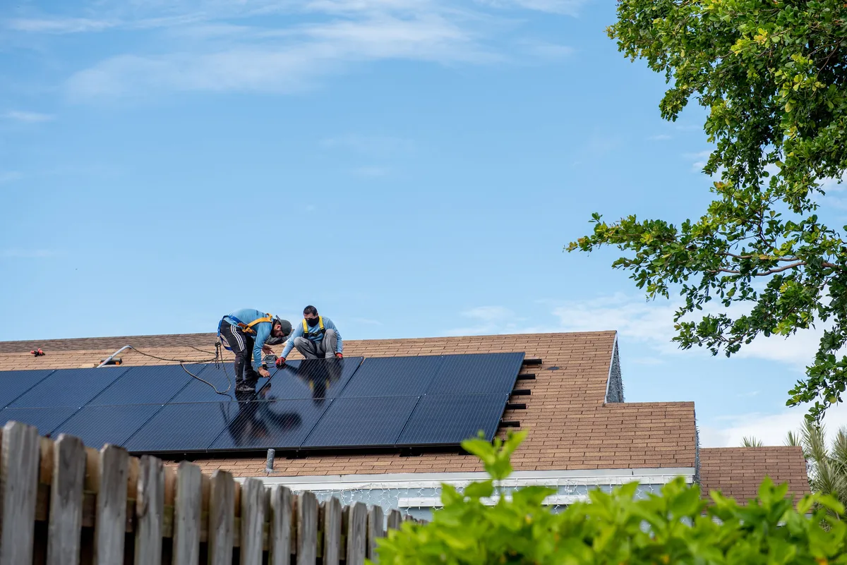 Two solar installers mounting black panels on an asphalt shingle roof under blue sky