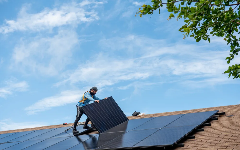 Solar installer lifting a black panel into position on a residential shingle roof