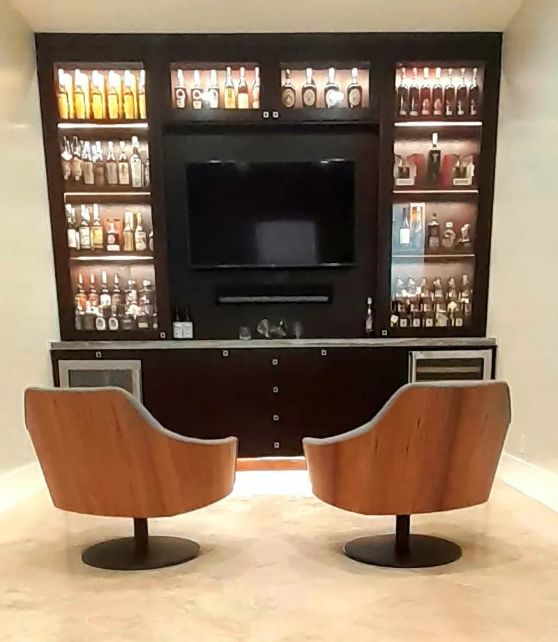Illuminated home bar with floor-to-ceiling bottle shelving, TV, and two swivel chairs
