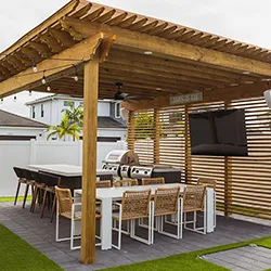Rustic wood pergola with string lights over long outdoor dining table and woven chairs, with mounted TV