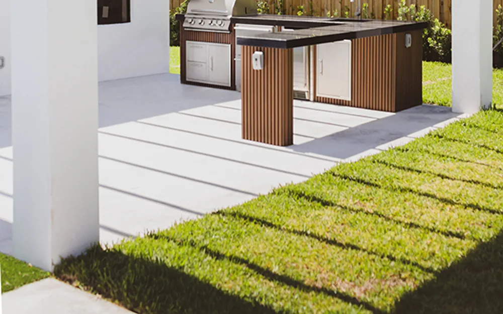 Outdoor kitchen island with stainless grill on a concrete patio beside lawn and white pergola columns